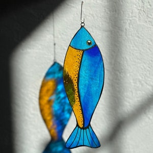 Peut inclure: Ornement de poisson en vitrail bleu et or, suspendu par un fil. Un deuxi&egrave;me poisson flou est visible en arri&egrave;re-plan. Le poisson a une nageoire caudale et est un objet d&eacute;coratif. Les couleurs sont vives et le design est attrayant.