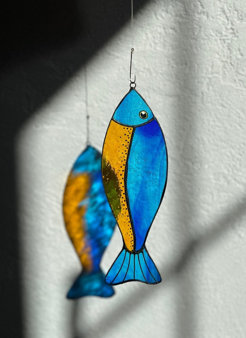 Peut inclure: Ornement de poisson en vitrail bleu et or, suspendu par un fil. Un deuxi&egrave;me poisson flou est visible en arri&egrave;re-plan. Le poisson a une nageoire caudale et est un objet d&eacute;coratif. Les couleurs sont vives et le design est attrayant.