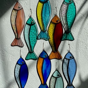Peut inclure: Une collection d'ornements de poissons en vitrail color&eacute;s, suspendus par un fil. Les poissons pr&eacute;sentent diverses couleurs, notamment le bleu, le vert, le jaune, l'orange et le rouge, avec des motifs d&eacute;taill&eacute;s d'&eacute;cailles et de nageoires. Id&eacute;al pour la d&eacute;coration sur le th&egrave;me marin.