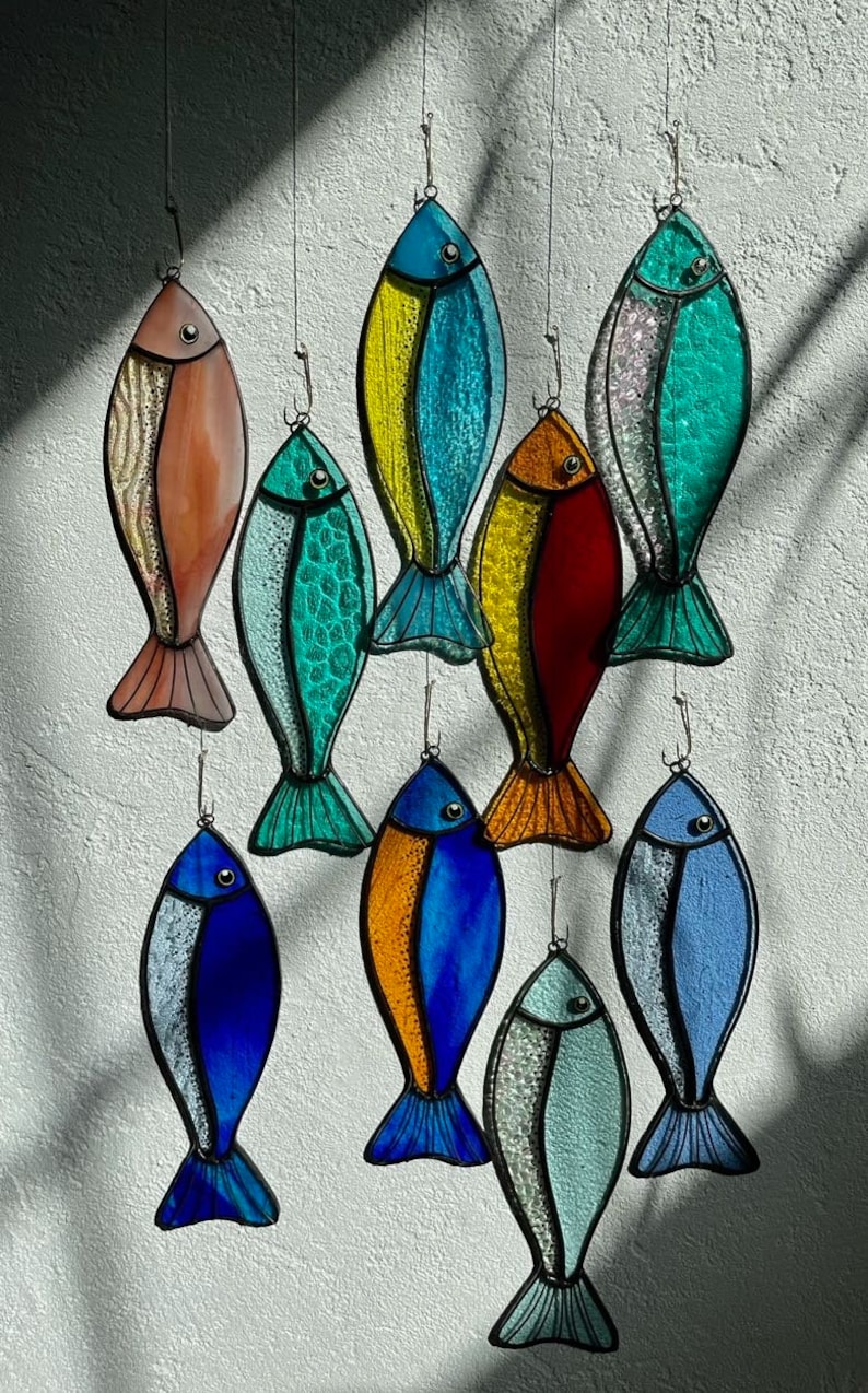 Peut inclure: Une collection d'ornements de poissons en vitrail color&eacute;s, suspendus par un fil. Les poissons pr&eacute;sentent diverses couleurs, notamment le bleu, le vert, le jaune, l'orange et le rouge, avec des motifs d&eacute;taill&eacute;s d'&eacute;cailles et de nageoires. Id&eacute;al pour la d&eacute;coration sur le th&egrave;me marin.