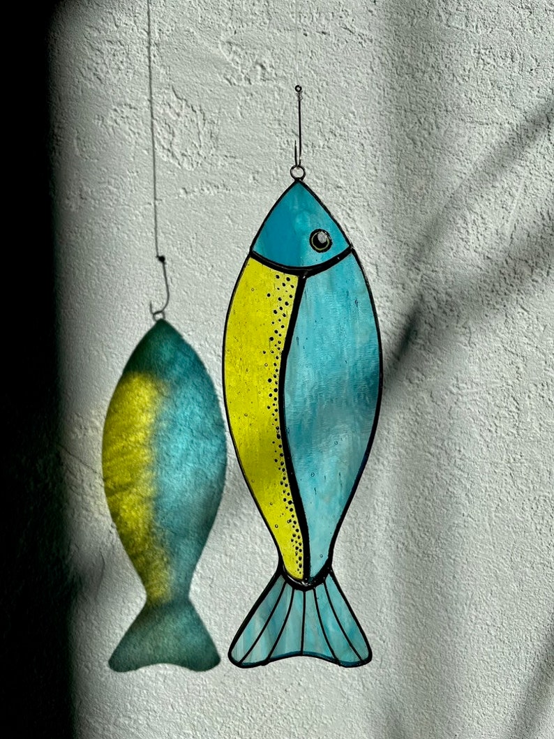 Peut inclure: Deux d&eacute;corations de poissons en vitrail suspendues devant un mur blanc textur&eacute;. Un poisson est jaune et bleu, l'autre est une ombre du premier. Les poissons ont des contours noirs et sont suspendus par de fins fils.