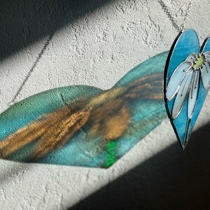 May include: A stained glass heart-shaped ornament with a blue background and a white flower with a green center. The ornament is hanging from a silver chain and casts a shadow on a white wall.