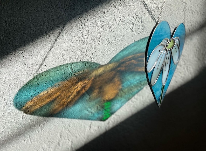 May include: A stained glass heart-shaped ornament with a blue background and a white flower with a green center. The ornament is hanging from a silver chain and casts a shadow on a white wall.