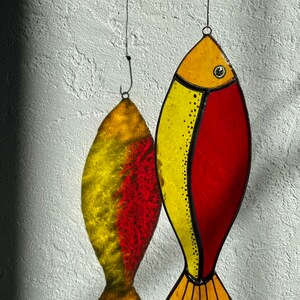 Peut inclure: Deux ornements de poisson en vitrail. L'un est jaune et rouge, avec un contour noir et un petit &oelig;il. L'autre est l'ombre du premier. Les deux sont suspendus &agrave; un fil fin contre un mur blanc textur&eacute;.