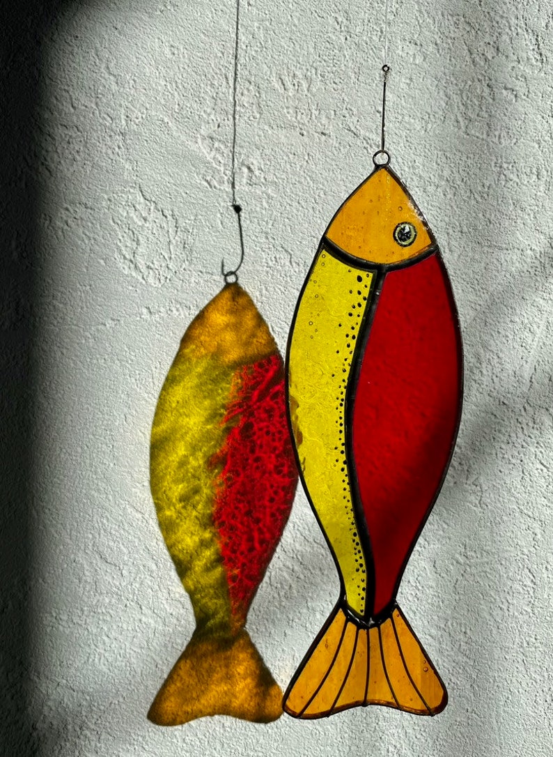 Peut inclure: Deux ornements de poisson en vitrail. L'un est jaune et rouge, avec un contour noir et un petit &oelig;il. L'autre est l'ombre du premier. Les deux sont suspendus &agrave; un fil fin contre un mur blanc textur&eacute;.