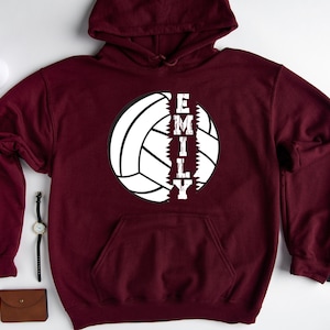 May include: Burgundy hoodie with a white volleyball graphic and the word "EMILY" printed vertically down the side of the ball.