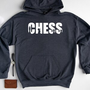 以下が含まれることがあります： 前面に白い「CHESS」という文字がプリントされたダークグレーのフード付きスウェットシャツ。 「CHESS」という文字はチェスの駒で構成されています。