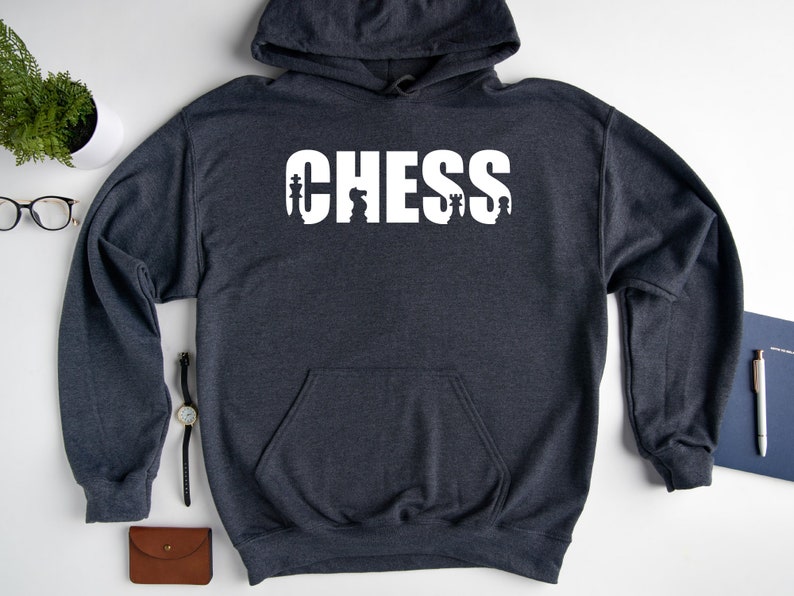 以下が含まれることがあります： 前面に白い「CHESS」という文字がプリントされたダークグレーのフード付きスウェットシャツ。 「CHESS」という文字はチェスの駒で構成されています。