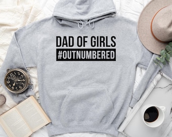 May include: A light gray hooded sweatshirt featuring the black text DAD OF GIRLS and the hashtag #OUTNUMBERED printed in white letters inside a black rectangular box on the center of the chest, including a front kangaroo pocket and long sleeves.