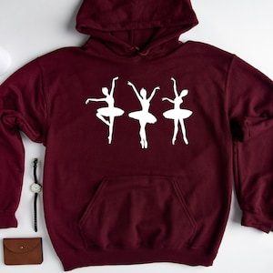 May include: Burgundy hoodie with a white silhouette of three ballerinas in a line. The ballerinas are wearing tutus and are in a dancing pose.