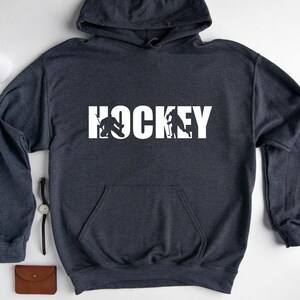 May include: A dark gray hooded sweatshirt with the word "HOCKEY" printed in white on the front. The word is made up of hockey players in silhouette.