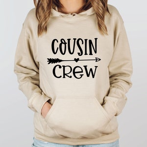 May include: A beige hoodie with a black graphic that says "COUSIN CREW" with an arrow and a heart.