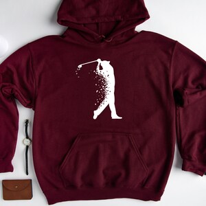 Op de afbeelding: Bordeauxkleurige hoodie met een wit silhouet van een golfer die een club zwaait. De golfer is gemaakt van verspreide witte stippen.