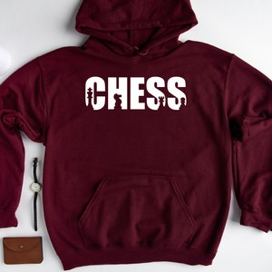 以下が含まれることがあります： ワインレッドのフード付きスウェットシャツに、白い文字で「CHESS」と書いてあり、文字の中にチェスの駒が組み込まれています。