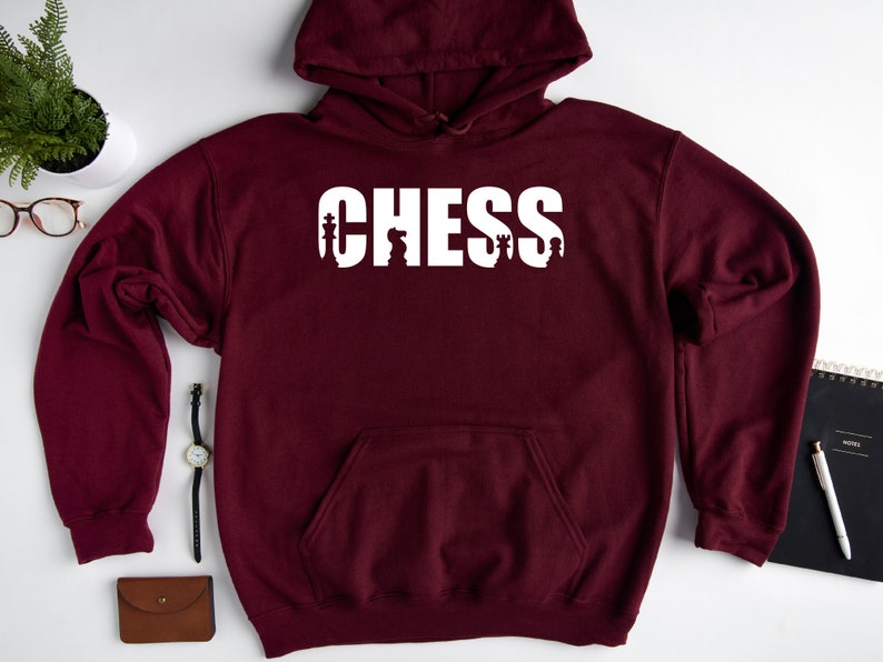 以下が含まれることがあります： ワインレッドのフード付きスウェットシャツに、白い文字で「CHESS」と書いてあり、文字の中にチェスの駒が組み込まれています。