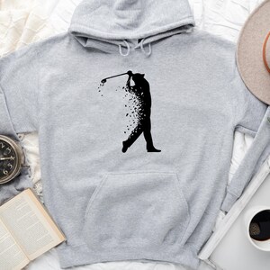 Op de afbeelding: Een grijze hoodie met een zwart silhouet van een golfer die een club zwaait. De golfer is gemaakt van verspreide zwarte vormen.
