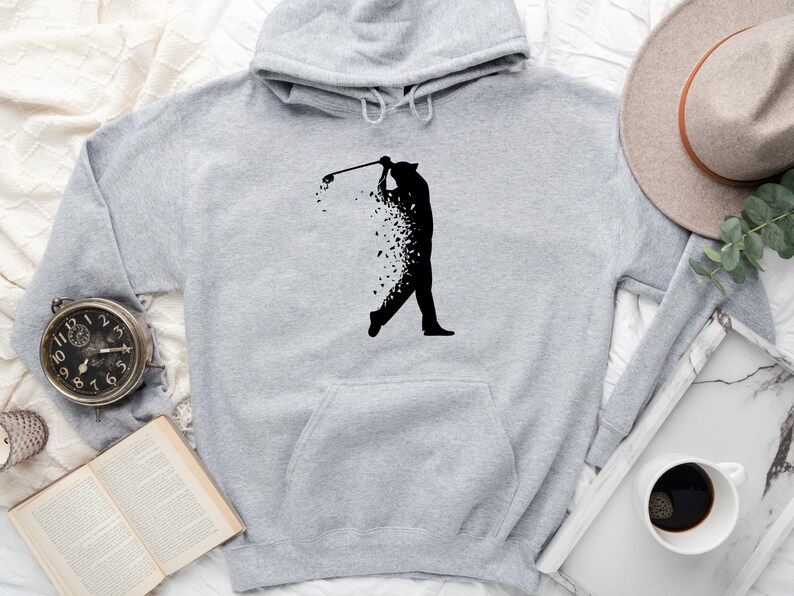 Op de afbeelding: Een grijze hoodie met een zwart silhouet van een golfer die een club zwaait. De golfer is gemaakt van verspreide zwarte vormen.