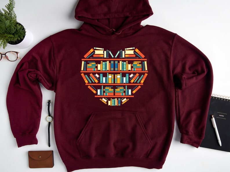 K&ouml;nnte beinhalten: Ein maroonfarbener Hoodie mit einem Aufdruck eines herzf&ouml;rmigen B&uuml;cherregals, das mit bunten B&uuml;chern gef&uuml;llt ist. Der Hoodie hat eine Kapuze mit Kordelzug und eine K&auml;ngurutasche.