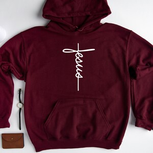 May include: Burgundy hooded sweatshirt with a white cross and the word "Jesus" written vertically on it.