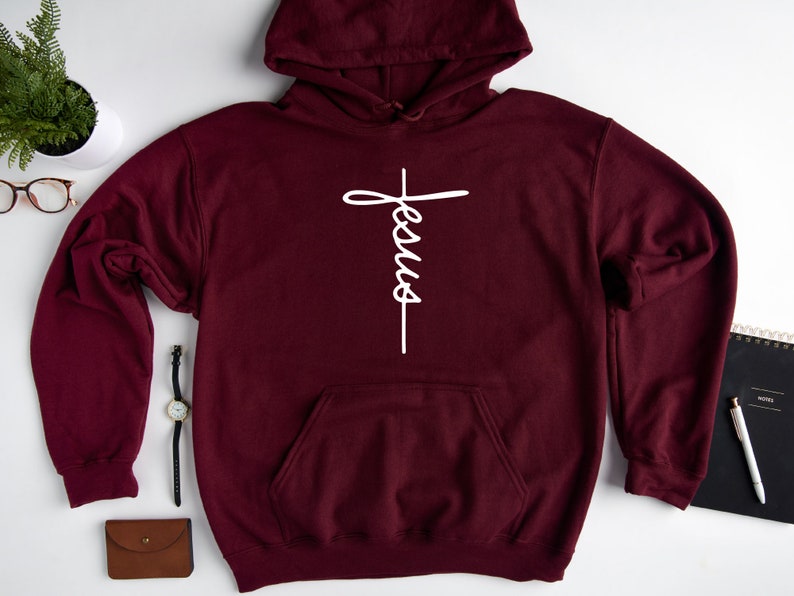 May include: Burgundy hooded sweatshirt with a white cross and the word "Jesus" written vertically on it.