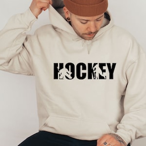 May include: A cream-colored hoodie with a black graphic of the word "HOCKEY" with hockey players silhouettes incorporated into the letters.