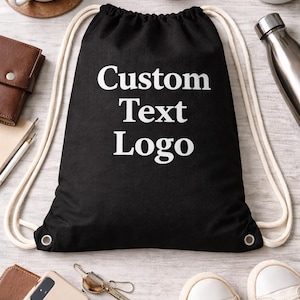 May include: Black drawstring bag with the text "Custom Text Logo" in white. The bag has white drawstrings and metal eyelets. Other items in the image include a cup of coffee, headphones, a plant, and a pair of white sneakers.
