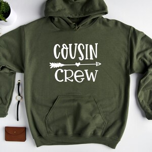 May include: A green hooded sweatshirt with the text "COUSIN CREW" in white lettering. An arrow with a heart in the middle is above the word "CREW".