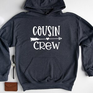 May include: A dark gray hooded sweatshirt with the text "COUSIN CREW" in white lettering. An arrow with a heart in the middle is above the word "CREW".
