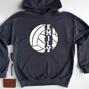 May include: A dark gray hooded sweatshirt with a white volleyball graphic and the name "EMILY" printed vertically down the center of the volleyball.