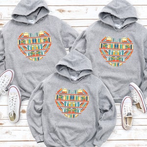 K&ouml;nnte beinhalten: Drei graue Kapuzenpullover mit einem bunten Herzdesign aus B&uuml;chern. Das Herzdesign enth&auml;lt den Text "Ich liebe B&uuml;cher".