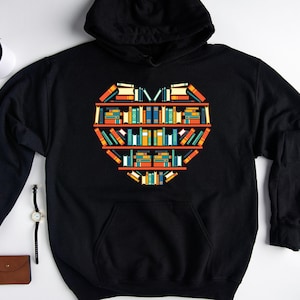 K&ouml;nnte beinhalten: Schwarzer Kapuzenpullover mit einem Grafikdruck eines Herzens aus B&uuml;chern in einem B&uuml;cherregal.