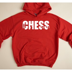 以下が含まれることがあります： チェスの駒が文字に組み込まれた「CHESS」という白いグラフィックが施された赤いフード付きスウェットシャツです。