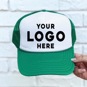 May include: A green and white trucker hat with the text "YOUR LOGO HERE" in black. The hat has a white front panel, a green brim, and a green mesh back. The hat is being held by a person.