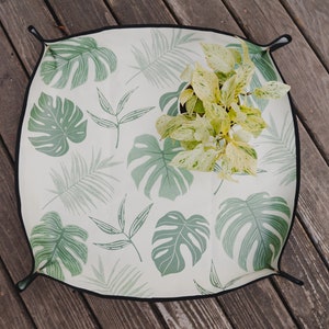 May include: A white picnic blanket with a green tropical leaf pattern. The blanket is folded and has a black border.