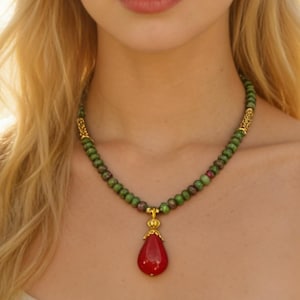 Könnte beinhalten: Halskette mit grünen und roten Perlen und goldenen Akzenten. Sie hat einen tropfenförmigen, roten Anhänger. Die Halskette ist ein Schmuckstück.