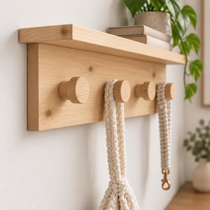 Peut inclure: Une étagère murale en bois clair composée d'une planche horizontale munie de quatre patères cylindriques. Une corde en macramé épaisse de couleur crème est suspendue à la deuxième patère, et un cordon assorti avec un fermoir métallique pend à la quatrième.