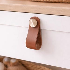 Könnte beinhalten: Eine weiße Keramik-Tasse mit schwarzem Henkel und dem Text "But First, Coffee" in fetter, schwarzer Schrift. Die Tasse steht vor einem verschwommenen Hintergrund, der eine gemütliche Umgebung suggeriert.