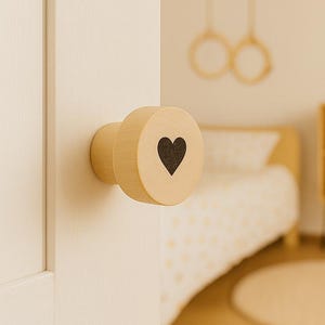 May include: A round, light-colored wooden door knob featuring a small, dark, solid heart shape printed in the center of its flat face. The knob is attached to a white door, with a blurred view of a room interior visible in the background.