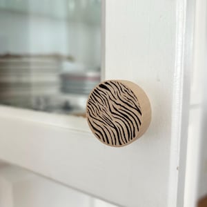 Peut inclure: Bouton de meuble en bois rond avec un motif zébré noir et beige. Le bouton est fixé à une porte d'armoire blanche. La porte est entrouverte, révélant des étagères en verre et de la vaisselle.