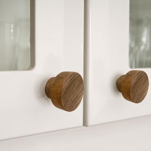May include: Two wooden cabinet knobs with a round, slightly raised top and a cylindrical base. The knobs are a dark brown colour and are mounted on a white cabinet door.