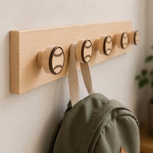 May include: A light-colored wooden wall-mounted rack featuring five circular knobs. Each knob is decorated with a dark brown engraved baseball pattern. A solid olive green backpack is hanging from the second knob on the left side of the rack.