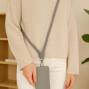 Puede incluir: Una funda de teléfono gris con un cordón trenzado a juego. El cordón se coloca alrededor del cuello y se sujeta a la funda del teléfono con un cierre metálico. La funda del teléfono es rectangular y tiene una superficie lisa.