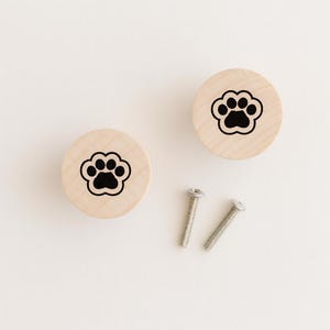 May include: Two wooden cabinet knobs featuring a black paw print design. The knobs are round and light-coloured, with two silver screws. Ideal for pet-themed decor.