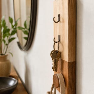 Peut inclure: Un support mural rectangulaire vertical en bois doté de deux crochets en métal foncé et d'une patère ronde en bois. La moitié supérieure est en bois clair, tandis que la moitié inférieure présente une finition en bois plus foncé.