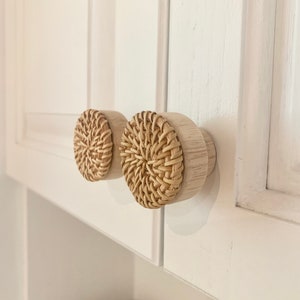 May include: Two round cabinet knobs with a natural woven rattan centre and light wood base. The knobs are attached to a white cabinet door. The knobs add a touch of natural texture to the cabinet.