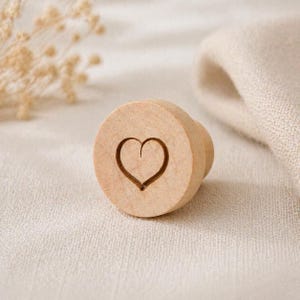 May include: A light-colored wooden bottle stopper with a heart-shaped design carved into the top. The stopper has a rounded top and a cylindrical base. The heart is a simple outline, adding a touch of elegance to the design.