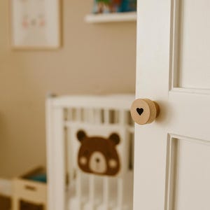 以下が含まれることがあります： 黒いハートの切り抜きが付いた木製のドアノブが白いドアに取り付けられています。背景には、茶色のクマのデザインの白いベビーベッドがあり、保育園のテーマを示唆しています。額入りのウサギのプリントと本棚も表示されています。