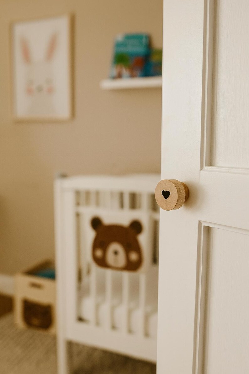 以下が含まれることがあります： 黒いハートの切り抜きが付いた木製のドアノブが白いドアに取り付けられています。背景には、茶色のクマのデザインの白いベビーベッドがあり、保育園のテーマを示唆しています。額入りのウサギのプリントと本棚も表示されています。