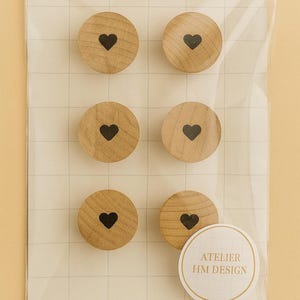 以下が含まれることがあります： 黒いハートのデザインが施された、6つの丸い木製マグネット。透明なプラスチックで包装されています。マグネットは明るい茶色で、木目が確認できます。「ATELIER HM DESIGN」と書かれたステッカーも付いています。