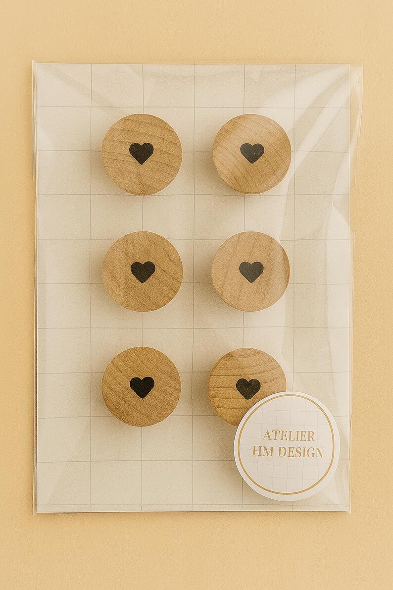 以下が含まれることがあります： 黒いハートのデザインが施された、6つの丸い木製マグネット。透明なプラスチックで包装されています。マグネットは明るい茶色で、木目が確認できます。「ATELIER HM DESIGN」と書かれたステッカーも付いています。